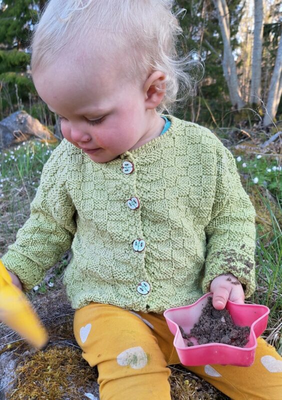 ett barn sitter på en sten i skogen med en spade och sandleksak i handen. Barnet har en grön stickad kofta på sig och gula byxor