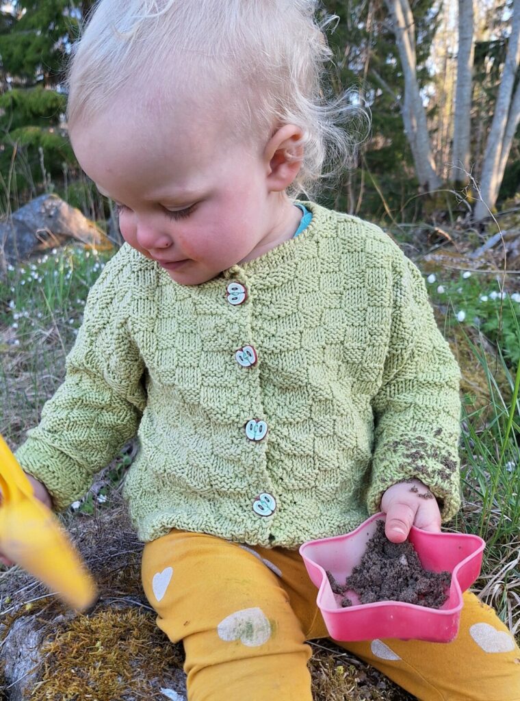 ett barn sitter på en sten i skogen med en spade och sandleksak i handen. Barnet har en grön stickad kofta på sig och gula byxor