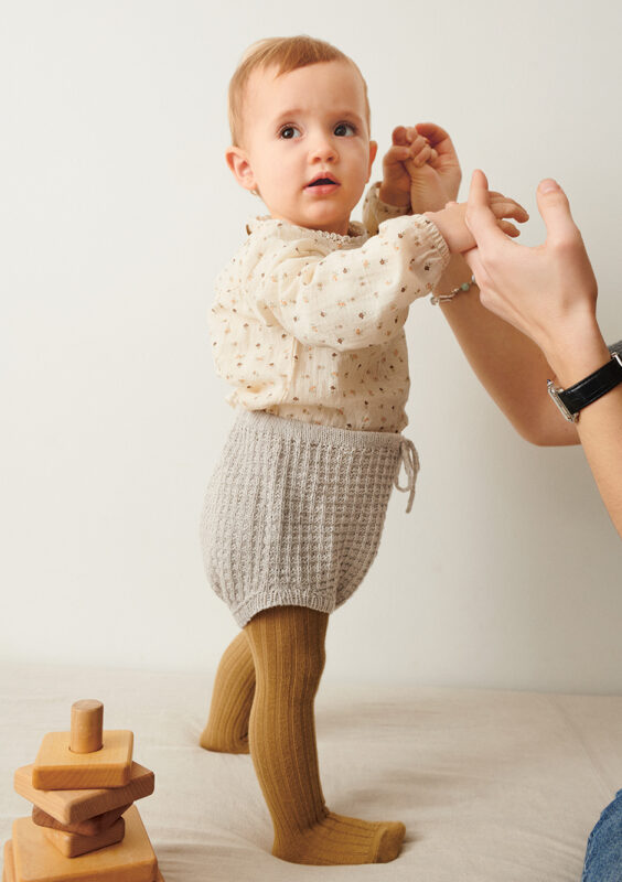 Liten bebis som står upp och bär grå Leo Bloomer Baby stickad i Sandnes Tynn Line Pärlgrå 3820 i mönster för barn från Sandnes Garn.