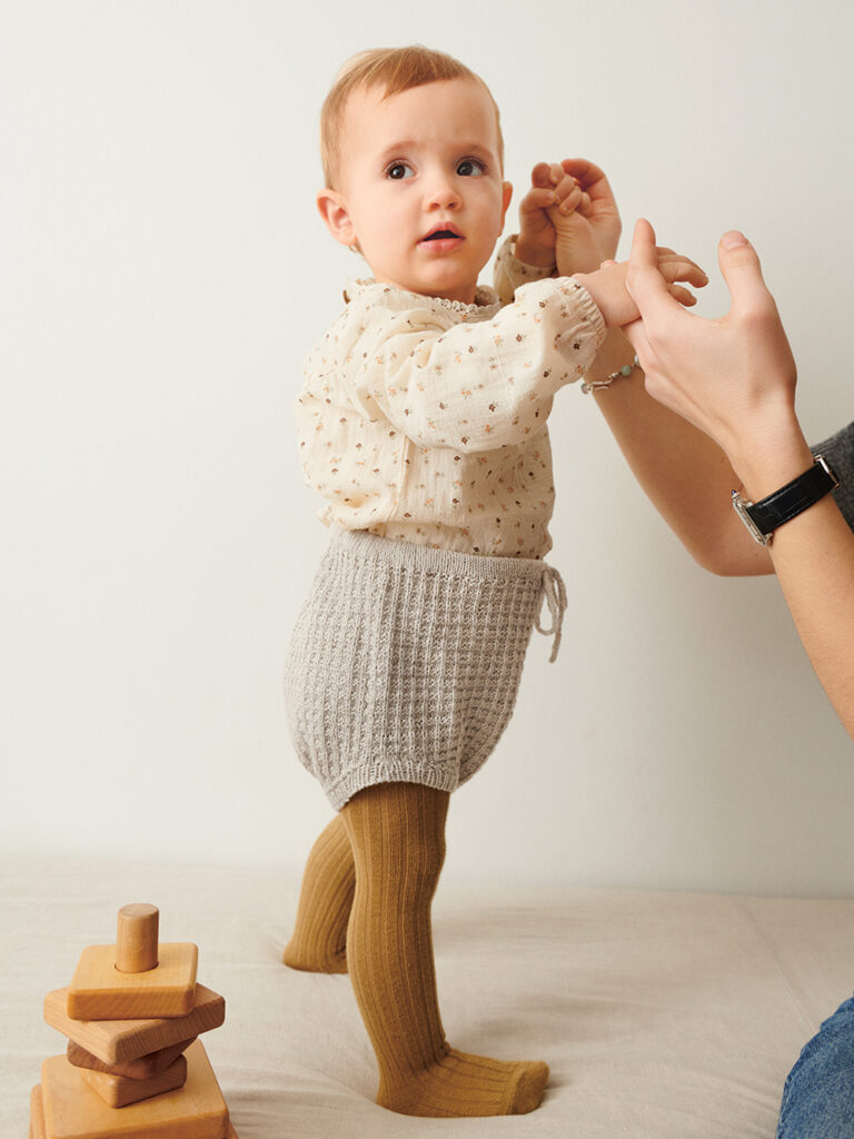 Liten bebis som står upp och bär grå Leo Bloomer Baby stickad i Sandnes Tynn Line Pärlgrå 3820 i mönster för barn från Sandnes Garn.