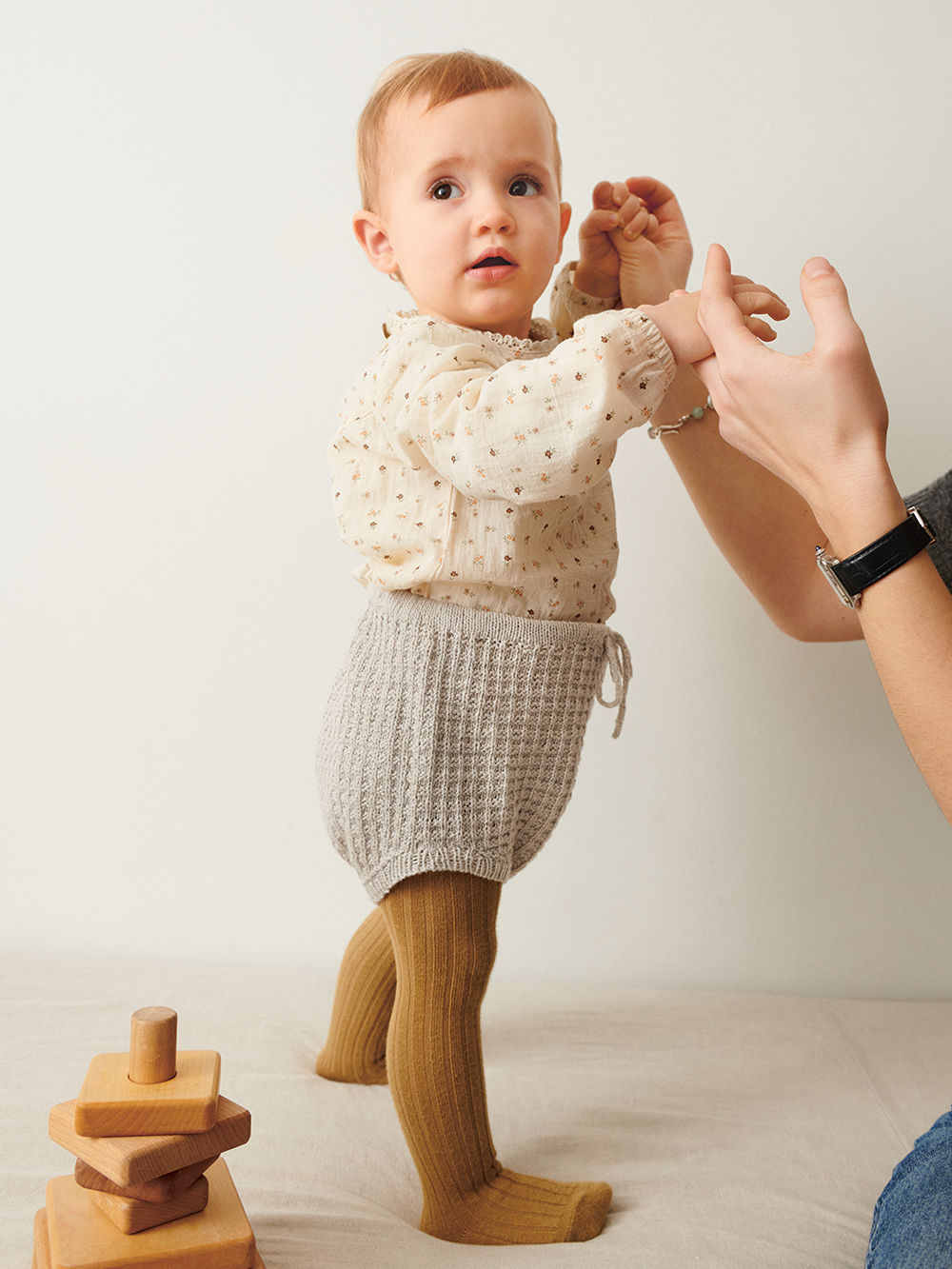 Liten bebis som står upp och bär grå Leo Bloomer Baby stickad i Sandnes Tynn Line Pärlgrå 3820 i mönster för barn från Sandnes Garn.