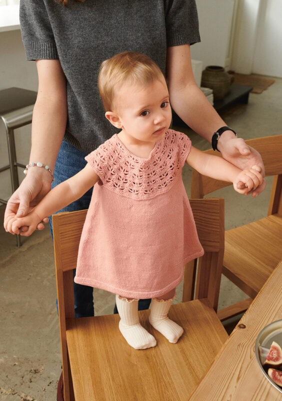 Liten bebis som står upp och bär rosa Indy Dress Baby stickad i Sandnes Mandarin Petit 4002 Ljus Persikoblomma i mönster för barn från Sandnes Garn.