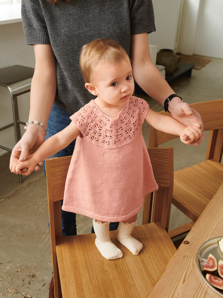 Liten bebis som står upp och bär rosa Indy Dress Baby stickad i Sandnes Mandarin Petit 4002 Ljus Persikoblomma i mönster för barn från Sandnes Garn.