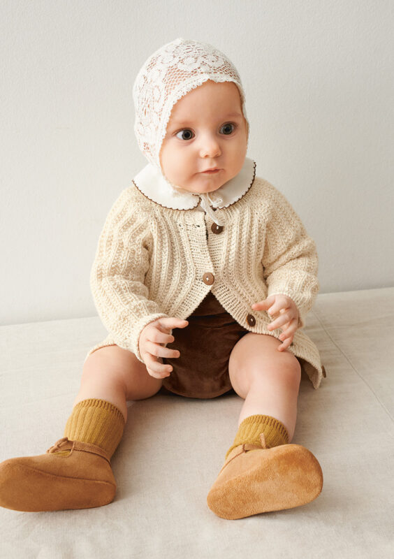 Bebis som sitter på golvet och bär vit Ellis Cardigan Baby stickad i Sandnes Mandarin Petit Natur 1012 i mönster för barn från Sandnes Garn.
