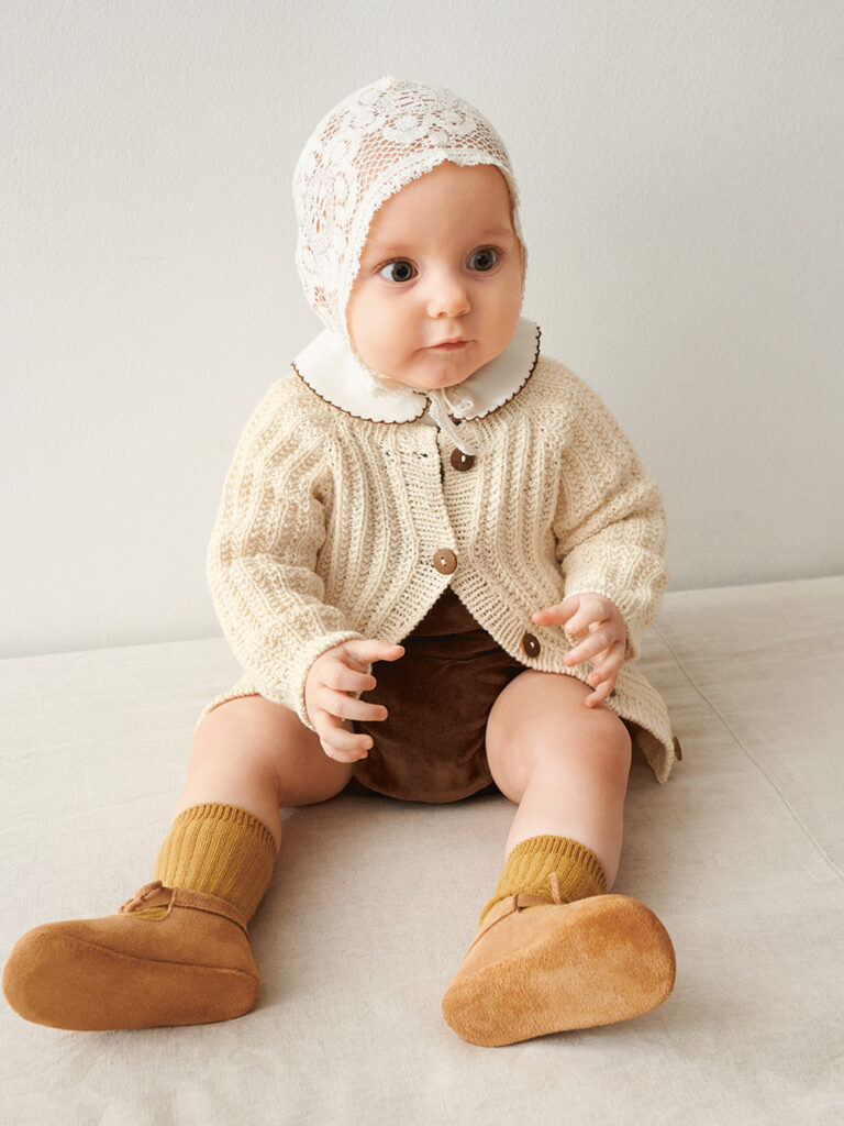 Bebis som sitter på golvet och bär vit Ellis Cardigan Baby stickad i Sandnes Mandarin Petit Natur 1012 i mönster för barn från Sandnes Garn.