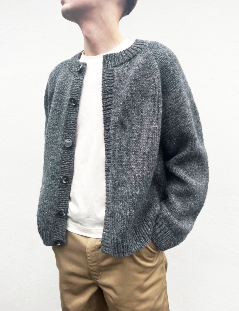 En man som bär vit t-shirt samt grå kofta med grå knappar stickat i mönster Foundation Cardigan Man från Le Knit.