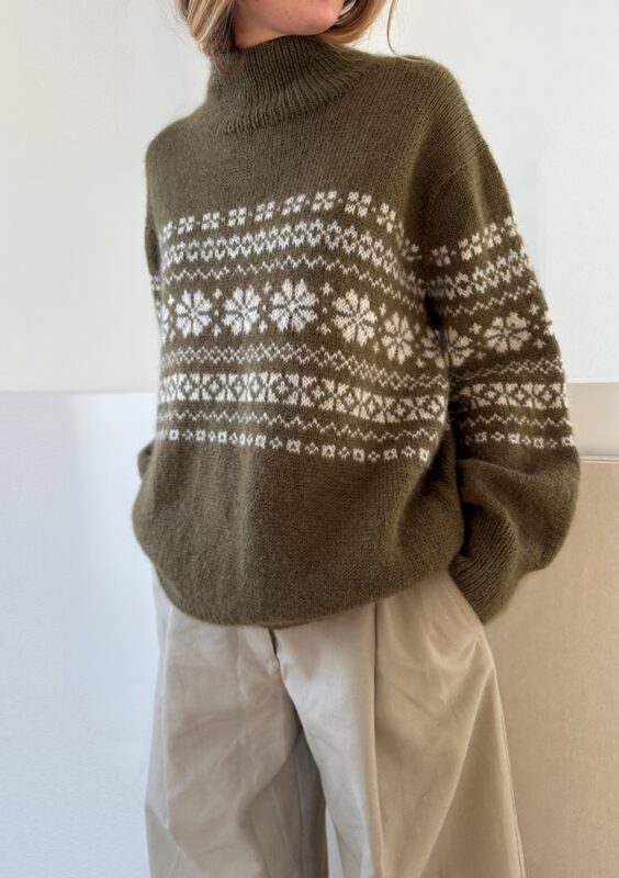 En kvinna som står upp och bär en mörkgrön tröja med vitt mönster stickad i mönster Harriet Sweater från Le Knit.