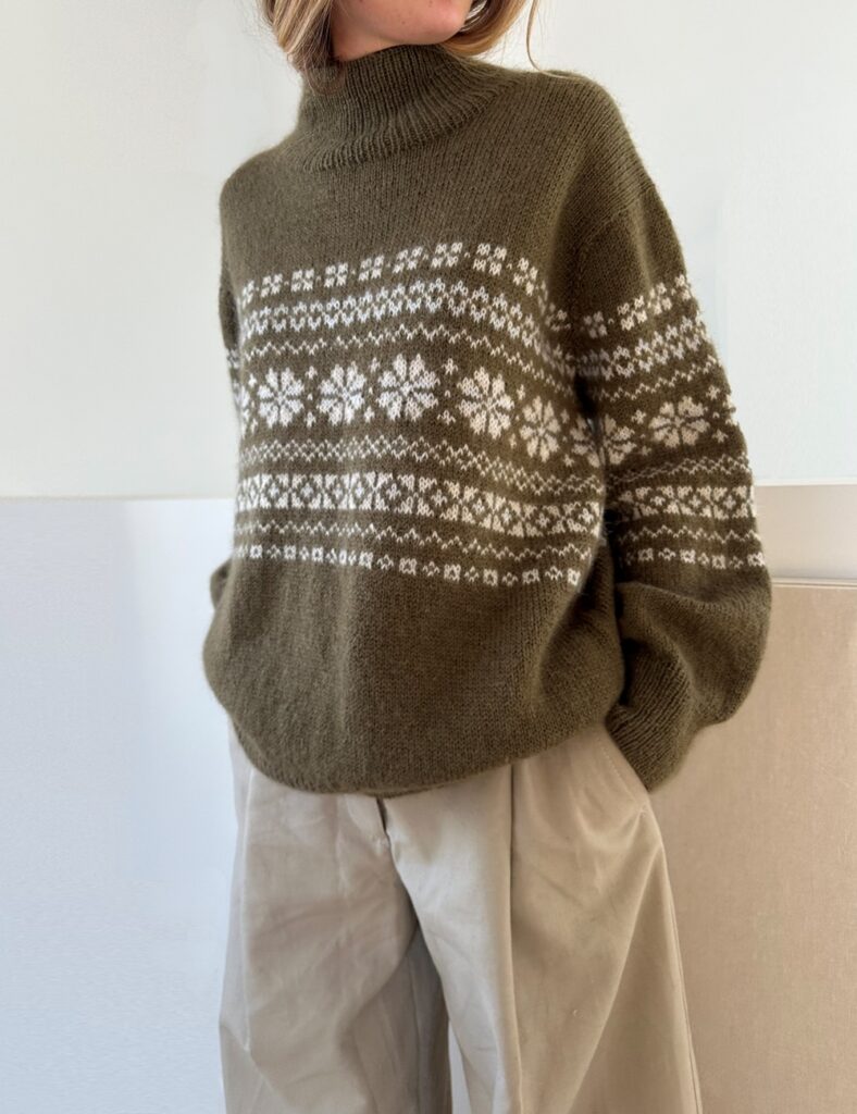 En kvinna som står upp och bär en mörkgrön tröja med vitt mönster stickad i mönster Harriet Sweater från Le Knit.