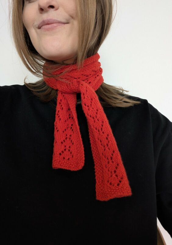 en kvinna har på sig en röd scarf i hålmönster virad runt halsen stickad i garnet sunday från sandnes