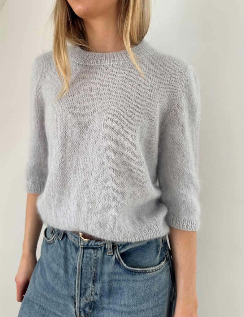 Kvinna med blonda lockar har på sig blå jeans och en stickad gråblå tröja med trekvartsärm, hon står med armarna längs med sidorna och ansiktet syns inte
