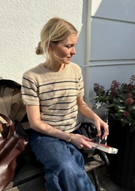 Kvinna sitter på en stol intill en husvägg i kvällssol och har en stickad beige tröja på sig med ränder i mörkblått. Hon håller en bok i handen.