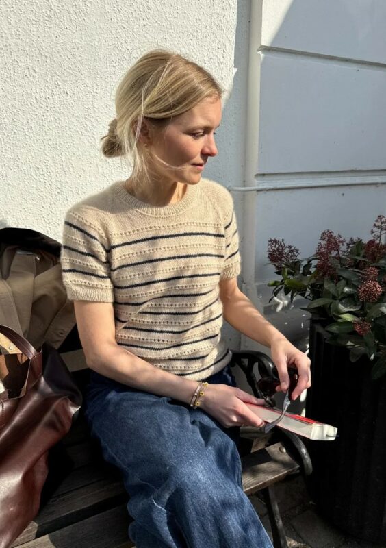 Kvinna sitter på en stol intill en husvägg i kvällssol och har en stickad beige tröja på sig med ränder i mörkblått. Hon håller en bok i handen.