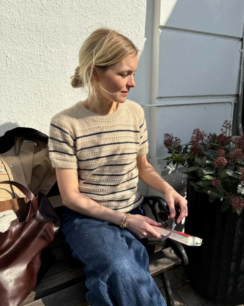 Kvinna sitter på en stol intill en husvägg i kvällssol och har en stickad beige tröja på sig med ränder i mörkblått. Hon håller en bok i handen.