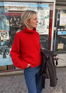 kvinna står utanför en butik med en hand i fickan och har på sig en klarröd stickad tröja med polokrage