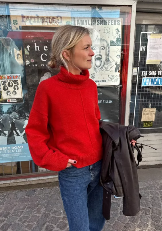 kvinna står utanför en butik med en hand i fickan och har på sig en klarröd stickad tröja med polokrage