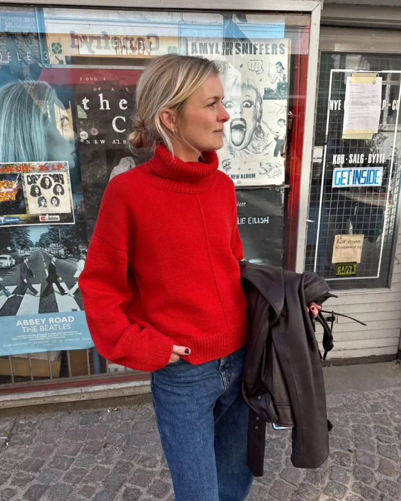 kvinna står utanför en butik med en hand i fickan och har på sig en klarröd stickad tröja med polokrage