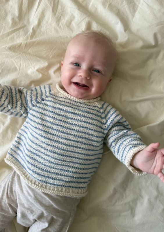 en bebis ligger på ett vitt skrynkligt lakan och har på sig en stickad blå tröja med vita ränder
