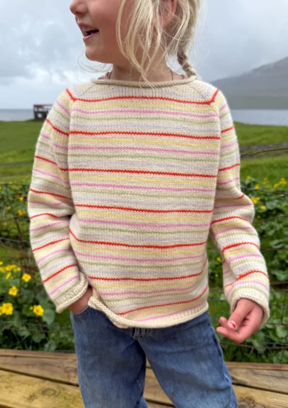 ett barn ute i naturen har på sig en stickad beige tröja med färgglada ränder och jeans.