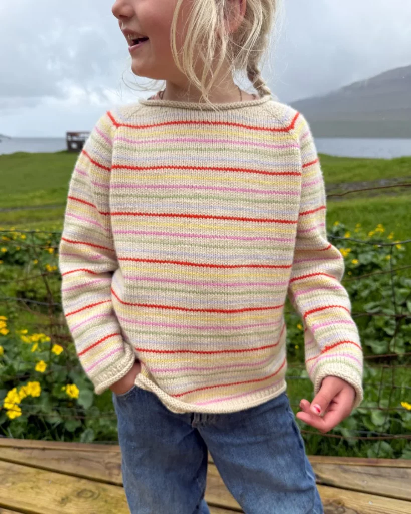 ett barn ute i naturen har på sig en stickad beige tröja med färgglada ränder och jeans.