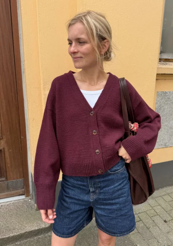 Kvinna som står mot en gul vägg klädd i Scarlet Cardigan från PetiteKnit.