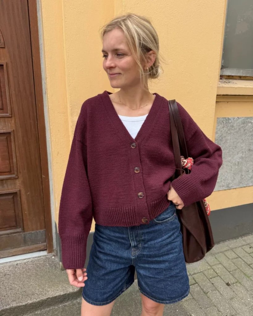 Kvinna som står mot en gul vägg klädd i Scarlet Cardigan från PetiteKnit.
