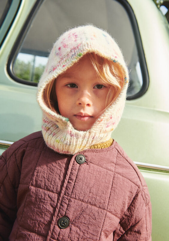 En flicka har på sig en balaclava och står framför en bil och tittar in i kameran