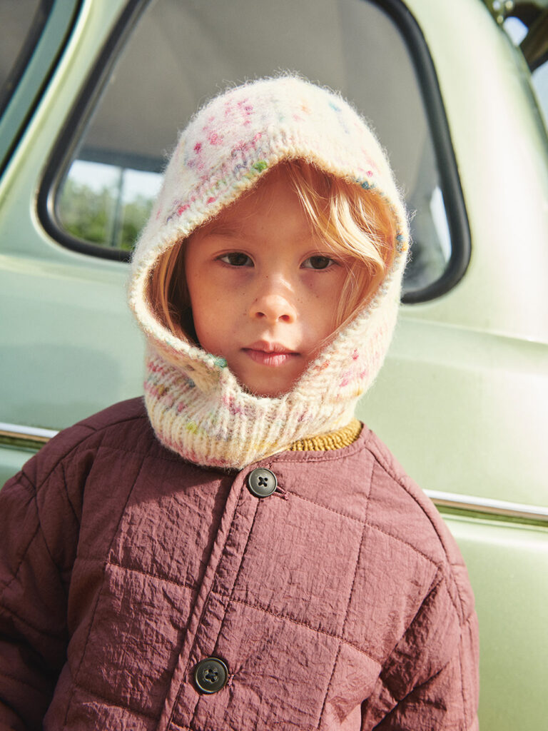 En flicka har på sig en balaclava och står framför en bil och tittar in i kameran