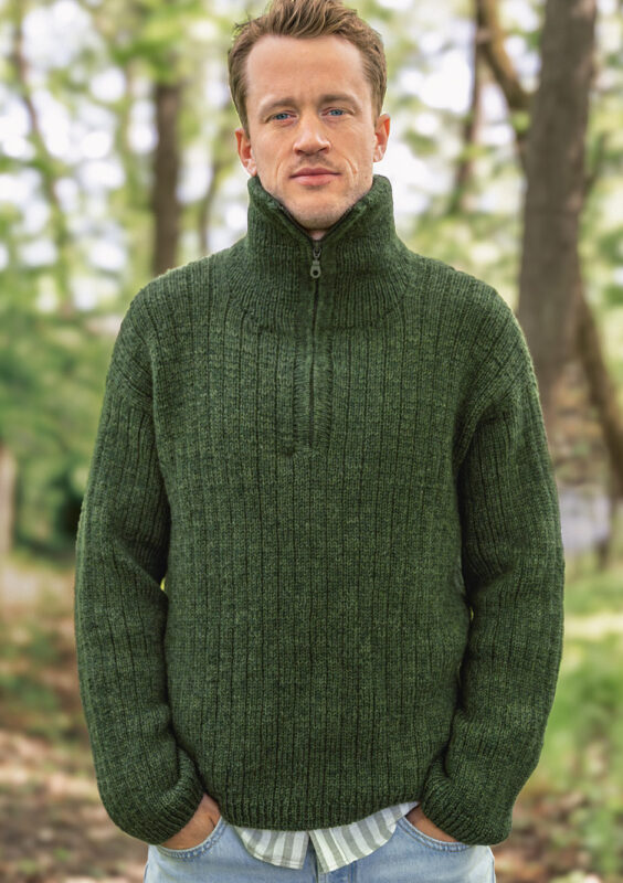 en man är i skogen och har händerna i fickorna, han har på sig en grön stickad tröja med polokrage
