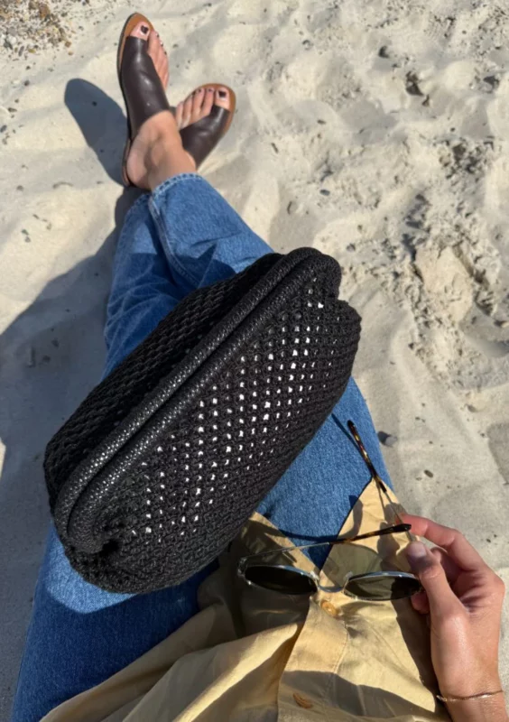en kvinna sitter med benen i kors på en sandstand och har en svart stickad clutch väska i knät och håller ett par solglasögon i handen