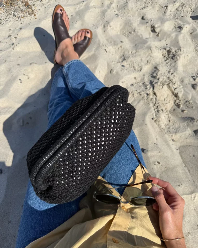 en kvinna sitter med benen i kors på en sandstand och har en svart stickad clutch väska i knät och håller ett par solglasögon i handen
